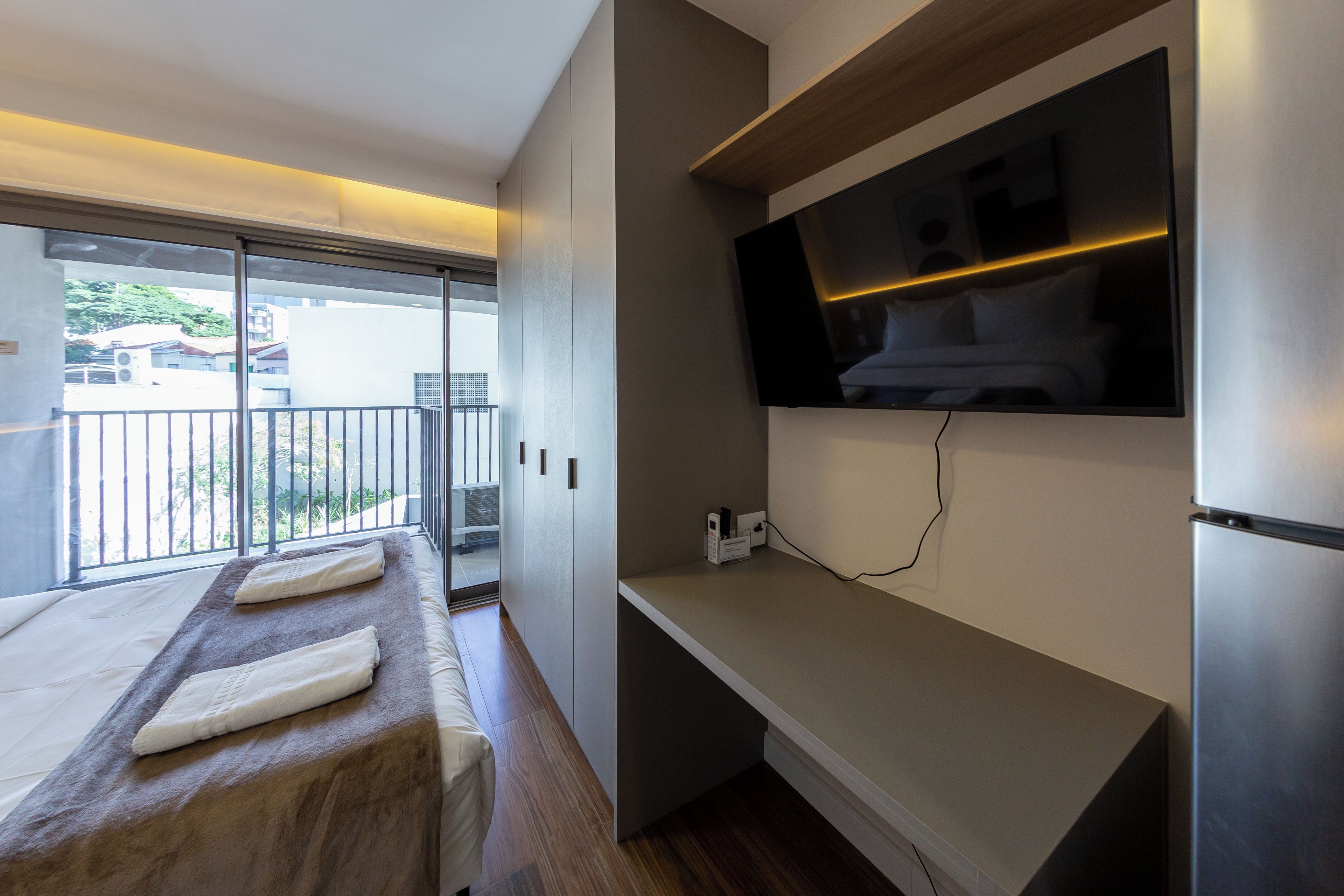 Et moderne hotelværelse med balkon, et fladskærms-tv ophængt på væggen og en træbænk med håndklæder.