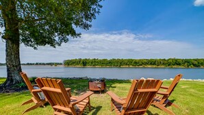 To træstole af Adirondack-typen vender ud mod en sø, med et træ og et bålsted i forgrunden.