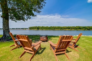Two wooden Adirondack chairs facing a lake with a tree and a fire pit in the foreground. - Riverfront Living (Clifton)
