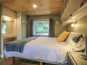 A bedroom with a bed, a bedside lamp, a window with curtains, a shelf with decorative items, and a mirror. - Orchard Lodge (Dover)