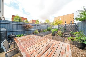 Un patio exterior con mesa y sillas de madera, rodeado por un jardín con diversas plantas y una valla azul.