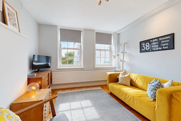 Ein modernes Wohnzimmer mit einem gelben Sofa, einem Fernseher und einem großen Fenster mit Blick auf Grünflächen.