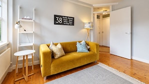 Una habitación con un sofá color mostaza, una mesita auxiliar blanca, una lámpara de pie, un radiador blanco y un cartel con el número 38.