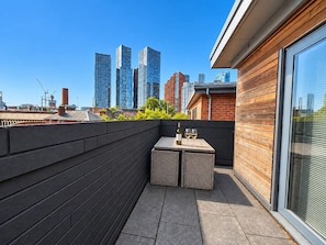 Eine Dachterrasse mit Tisch und Stühlen, einer Weinflasche und Blick auf hohe Gebäude.