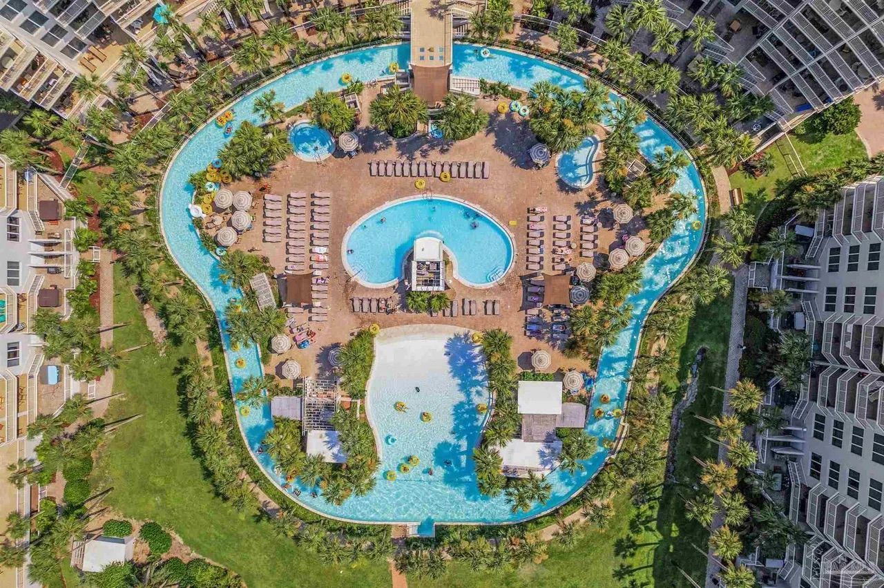 An aerial view of a resort with a central swimming pool surrounded by palm trees and sun loungers.