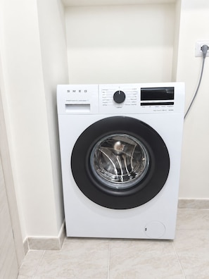 A white front-loading washing machine with a control panel, a power cord, and a tiled floor. - Pinuccia and Totore Sweet Home (Napoli)