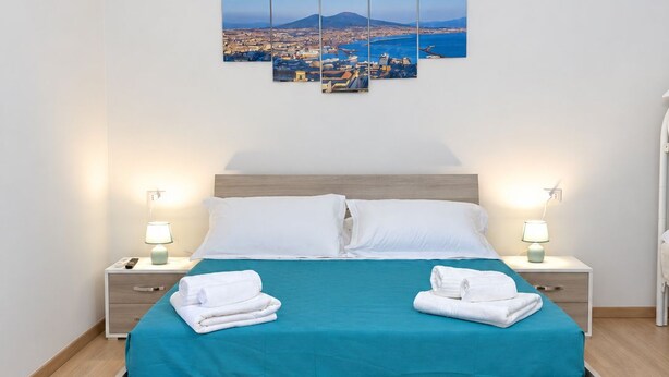 A hotel room with a bed, two nightstands, a wall-mounted artwork, and a view of a coastal town. - Vrbo Property (Napoli)