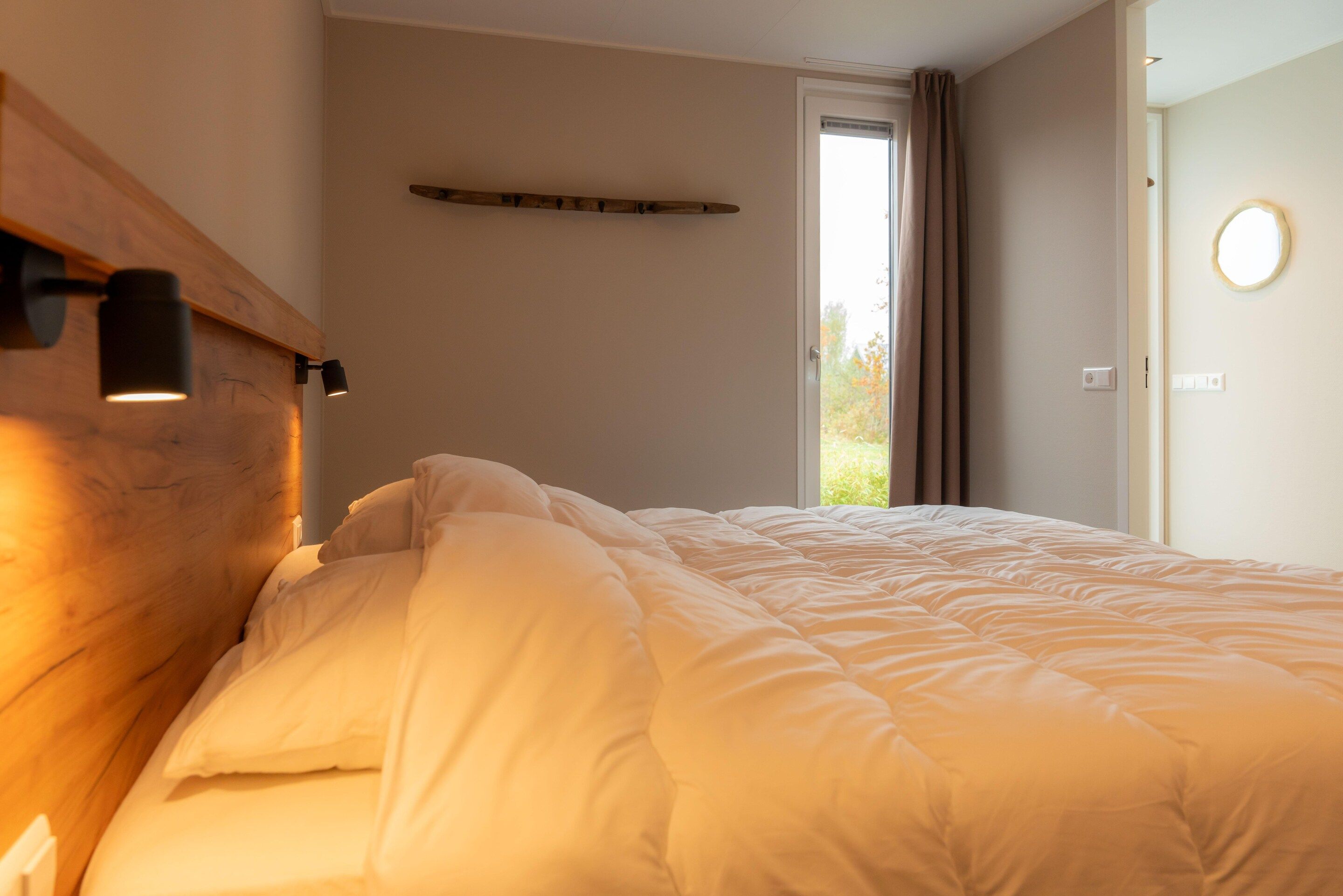Quarto com cama, cabeceira de madeira, luminária de cabeceira, janela com cortinas e porta com espelho redondo.