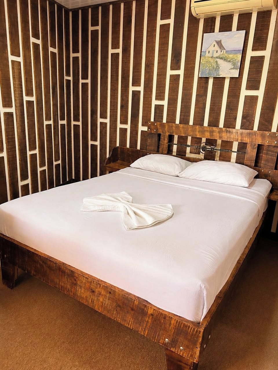 Una cama bien hecha con sábana y toalla blancos, cabecera de madera, y pared con listones verticales.
