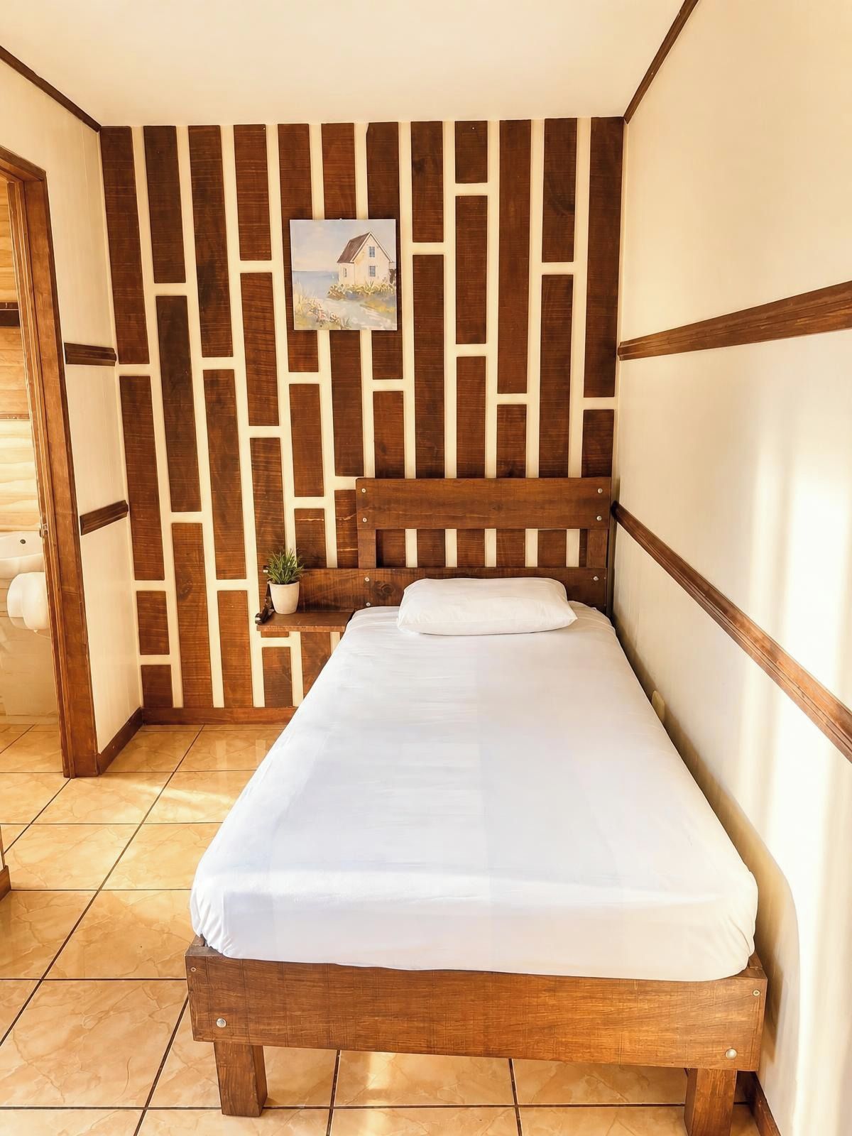 Una cama individual con estructura de madera y ropa de cama blanca en una habitación con pared de listones de madera y una pequeña planta en maceta.