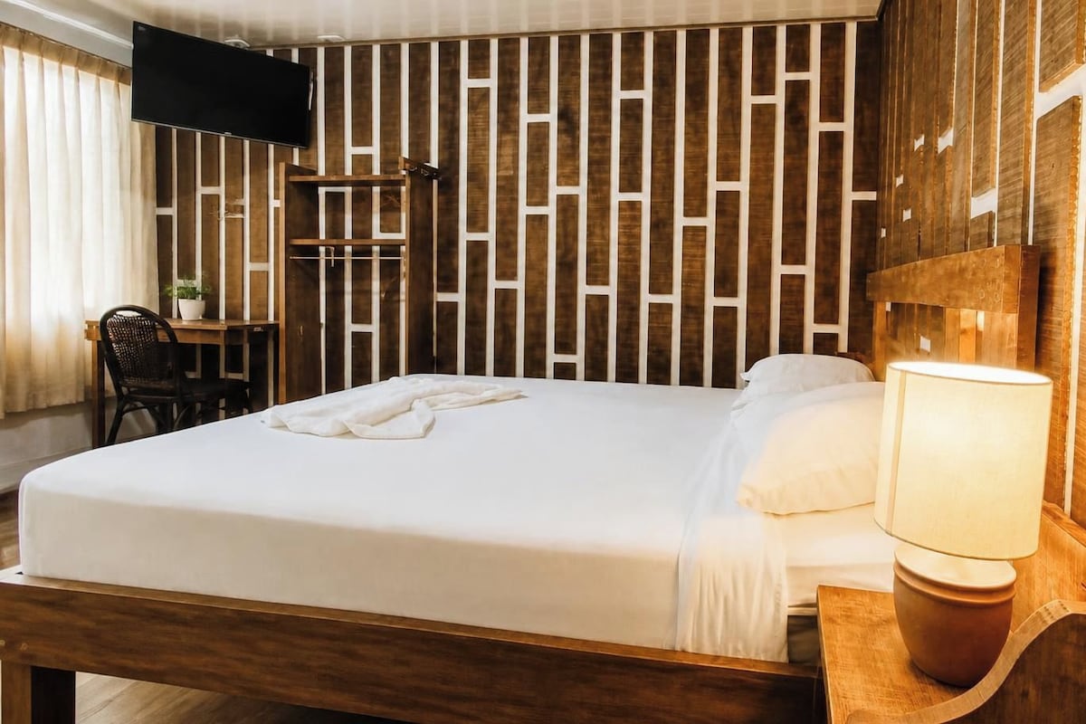 A hotel room with a bed, a desk, a chair, a TV mounted on the wall, and a wooden headboard.