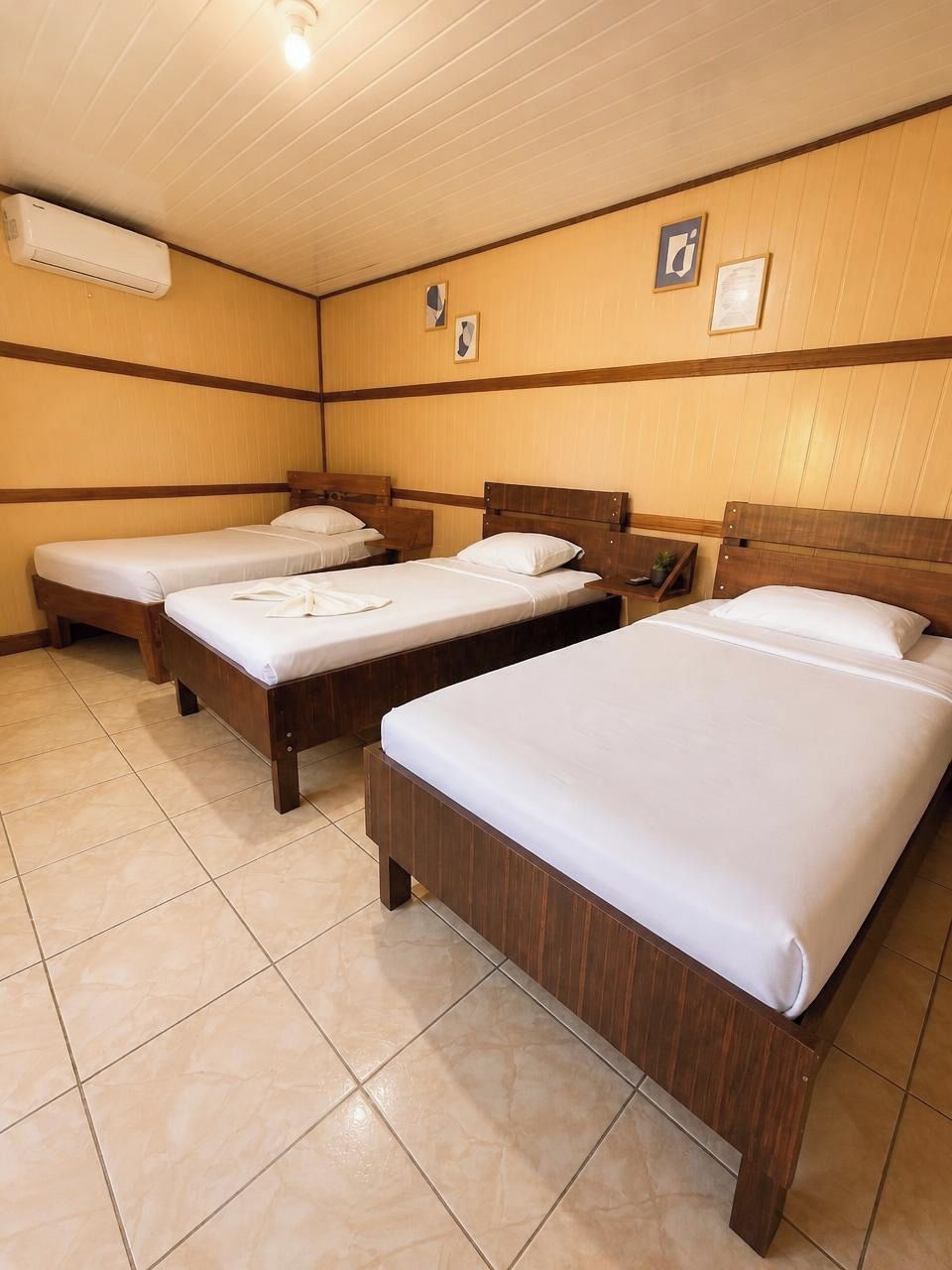 Une chambre d’hôtel avec trois lits en bois, un sol carrelé et un plafond muni d’un luminaire.