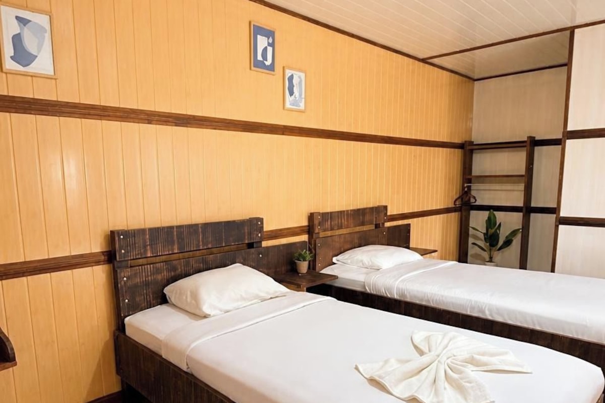 A hotel room with two wooden beds, a wooden headboard, a small bedside table with a plant, and a window with a view.