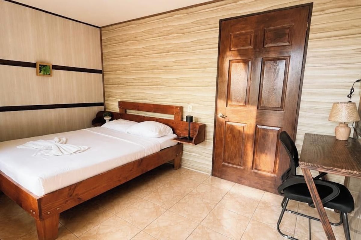 A bedroom with a bed, a desk, a chair, a wooden door, and a wall-mounted fan.