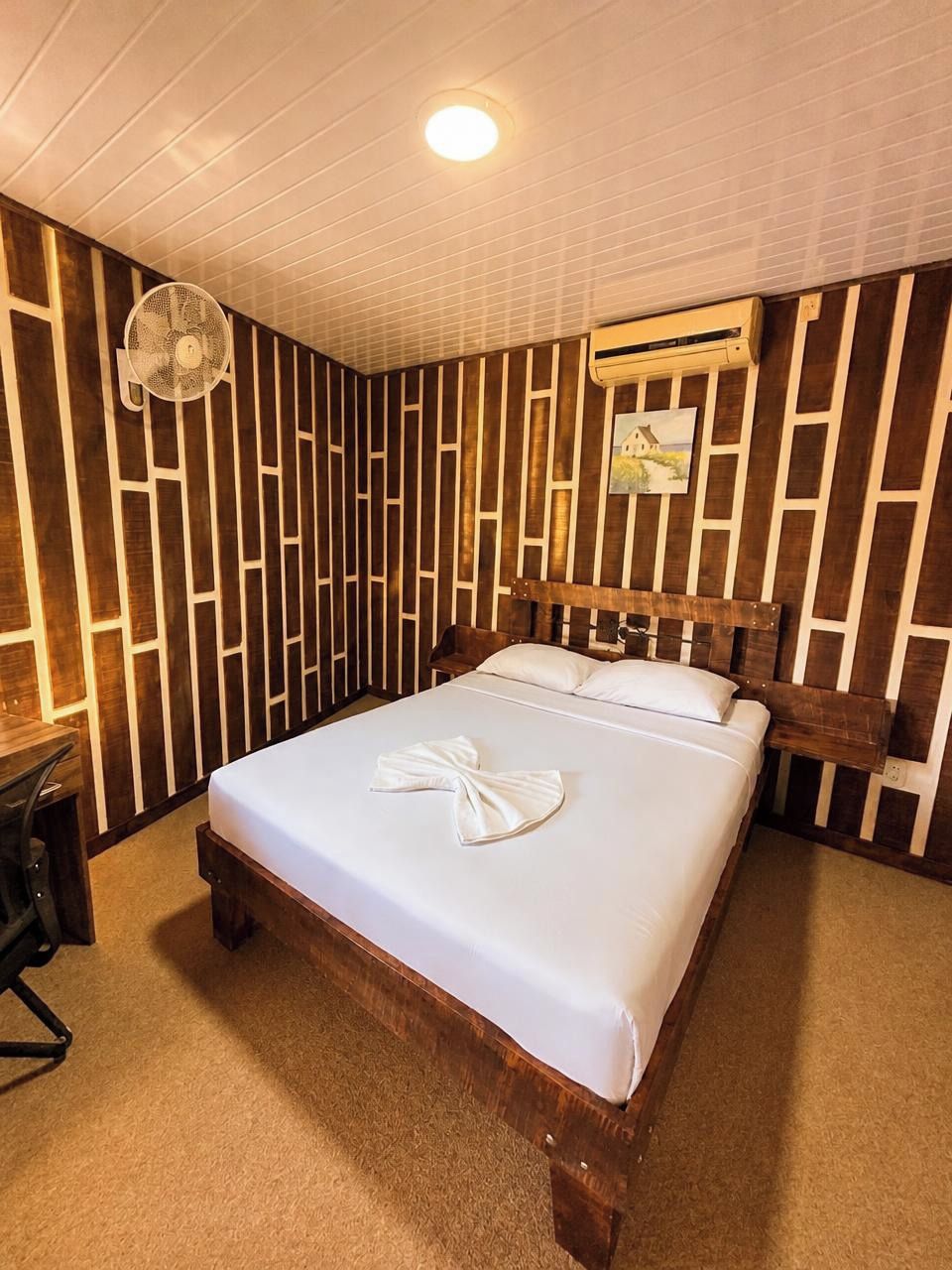 Habitación de hotel con cama de madera, escritorio, ventilador de pared y aire acondicionado.