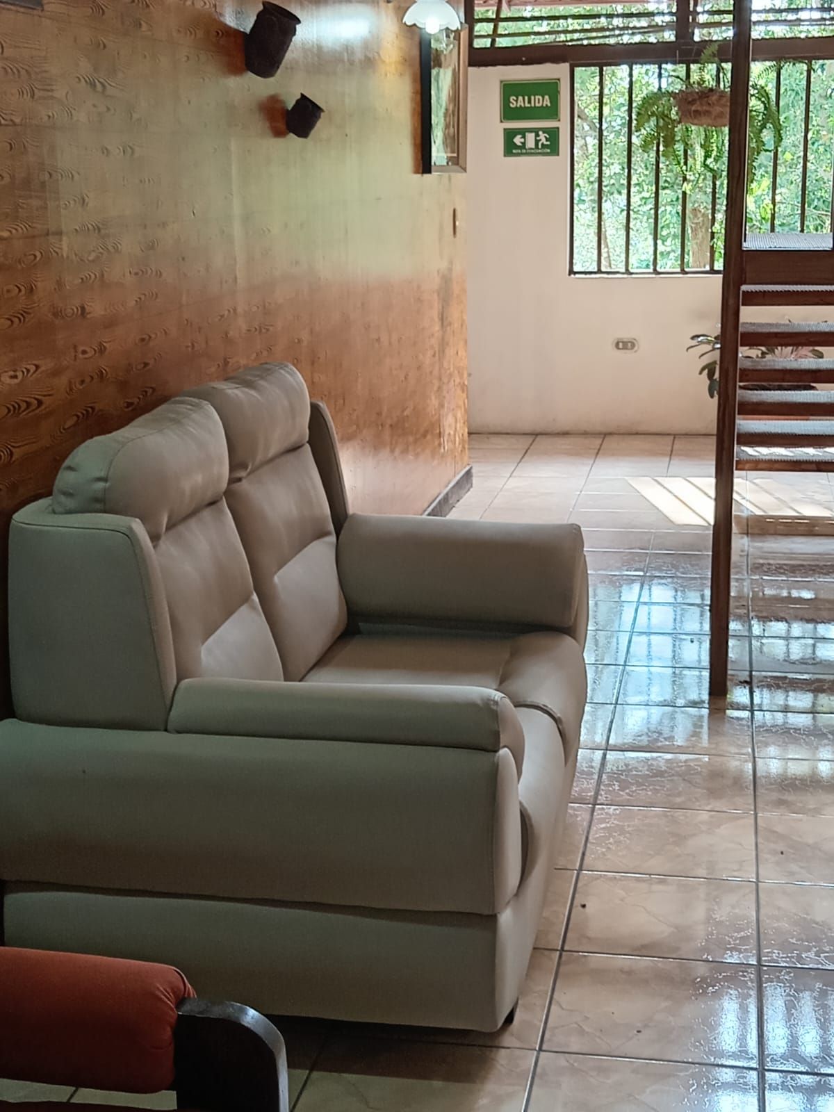 A beige sofa in a room with a tiled floor, a wooden wall, and a window with a view of greenery.