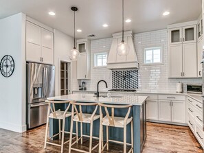 Une cuisine moderne avec des meubles blancs, un réfrigérateur en acier inoxydable, un grand îlot central doté de tabourets, des luminaires suspendus et un mur de fond en briques.