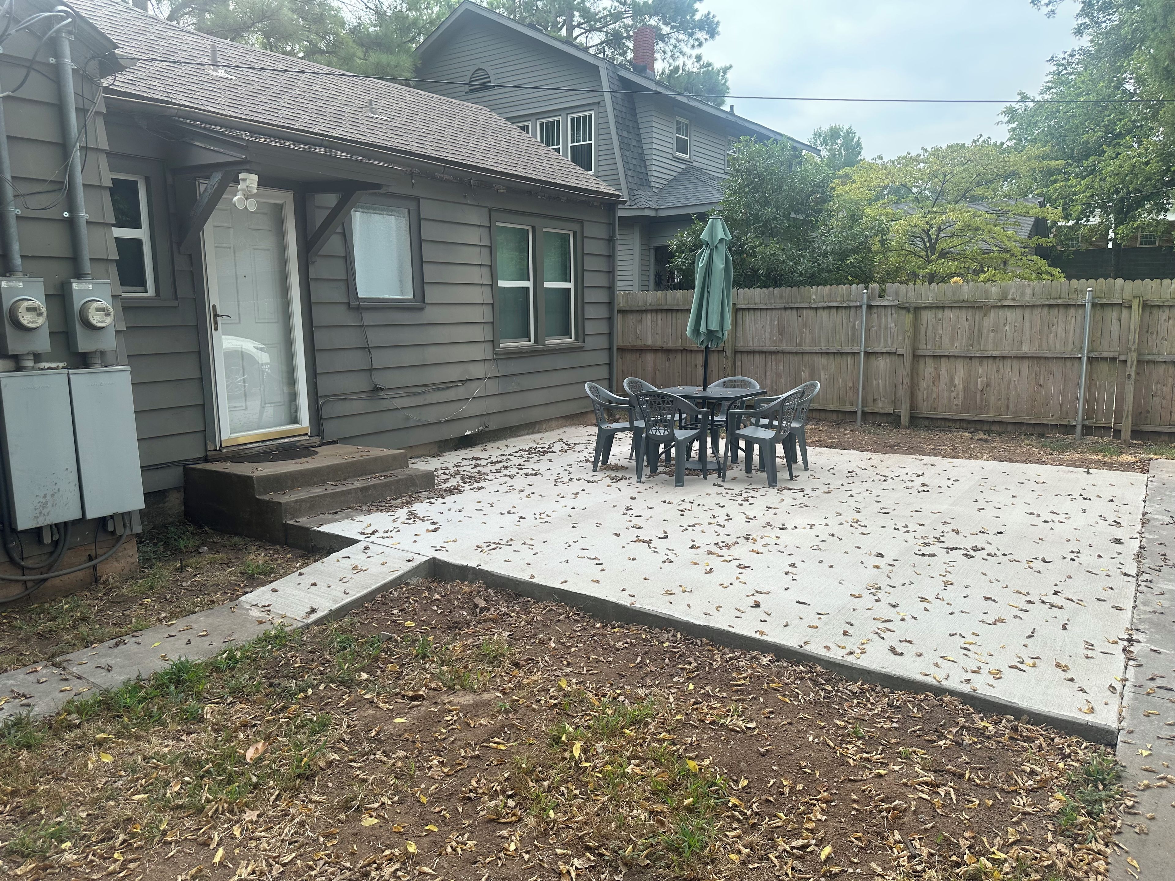 Un jardin à l’arrière d’une maison, avec une terrasse en béton, une clôture en bois et une table entourée de chaises.