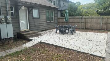 Un jardin à l’arrière d’une maison, avec une terrasse en béton, une clôture en bois et une table entourée de chaises.