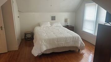 Une chambre à coucher comprenant un lit, une table de chevet, un ventilateur de plafond, une fenêtre avec des stores et une porte.