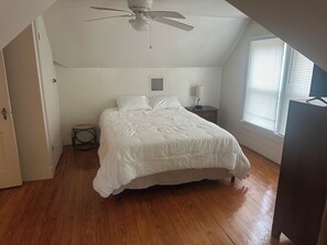 A bedroom with a bed, a nightstand, a ceiling fan, a window with blinds, and a door.
