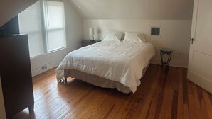 Et soveværelse med skråvæg, seng, natbord, lampe, et vindue med persienner og en loftsvifte.