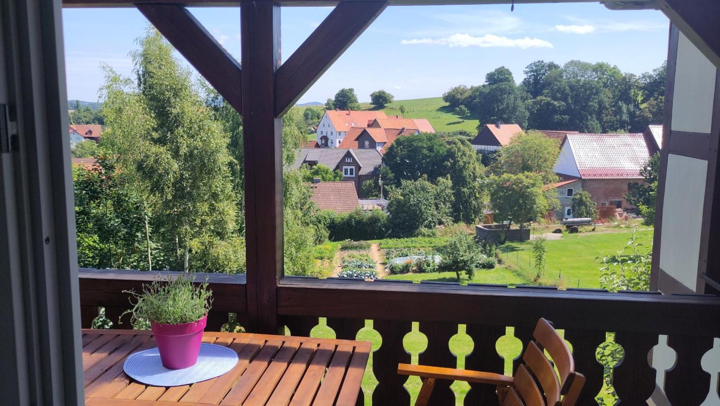 Eine Holzterrasse mit Tisch und Stühlen, einem Blumentopf und Blick auf eine ländliche Landschaft mit Häusern und viel Grün.