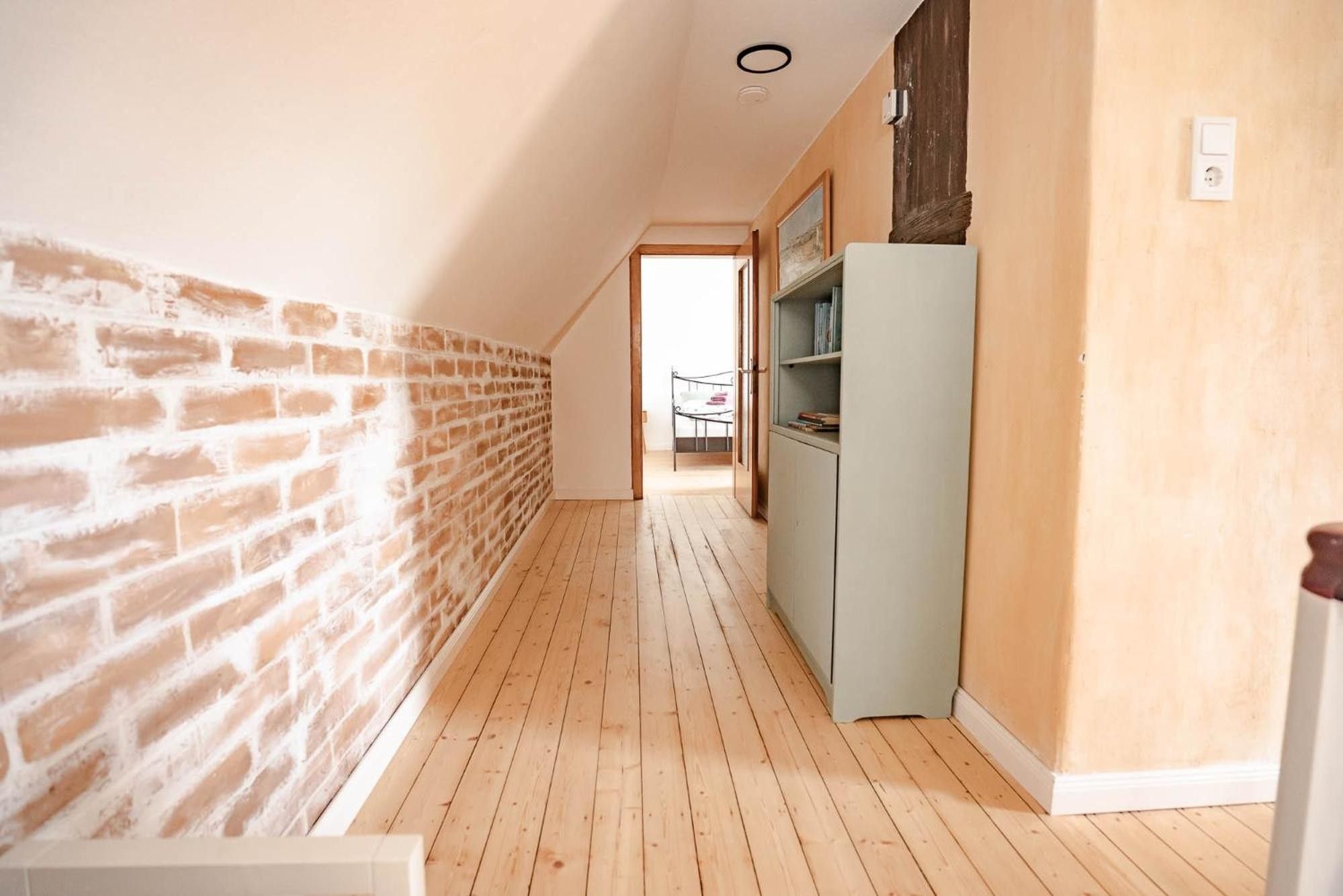 Ein schmaler Flur mit einer Backsteinwand und einer weißen Wand, der zu einem Schlafzimmer mit Bett und Kleiderschrank führt.