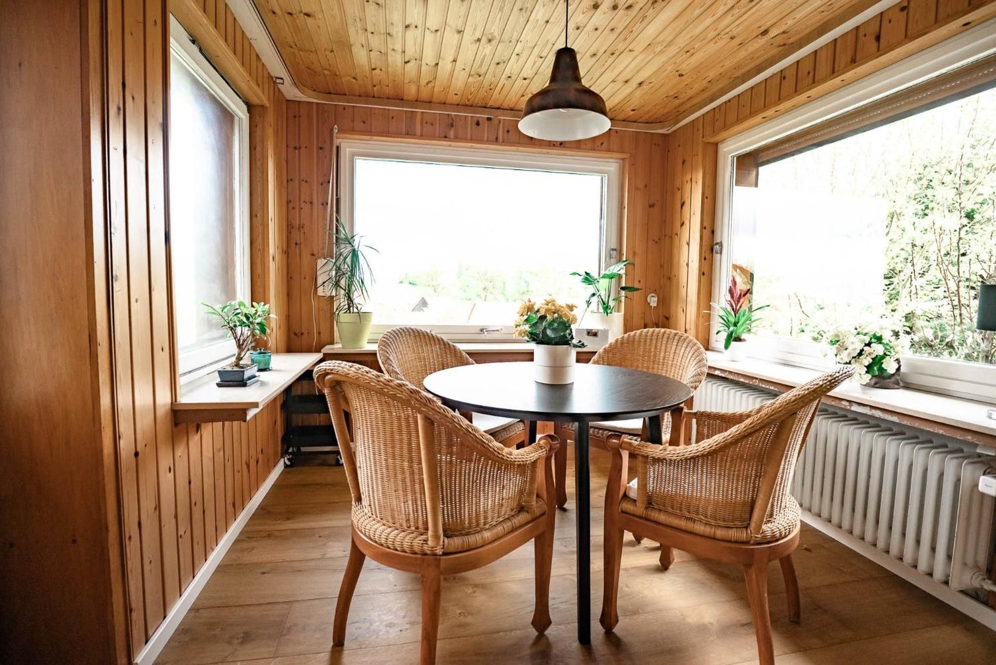 Ein Zimmer mit Holzwänden, einem runden Tisch und vier Korbstühlen, großen Fenstern und Topfpflanzen.