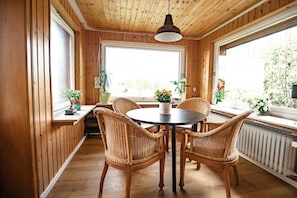 Ein Zimmer mit Holzwänden, einem runden Tisch und vier Korbstühlen, großen Fenstern und Topfpflanzen.