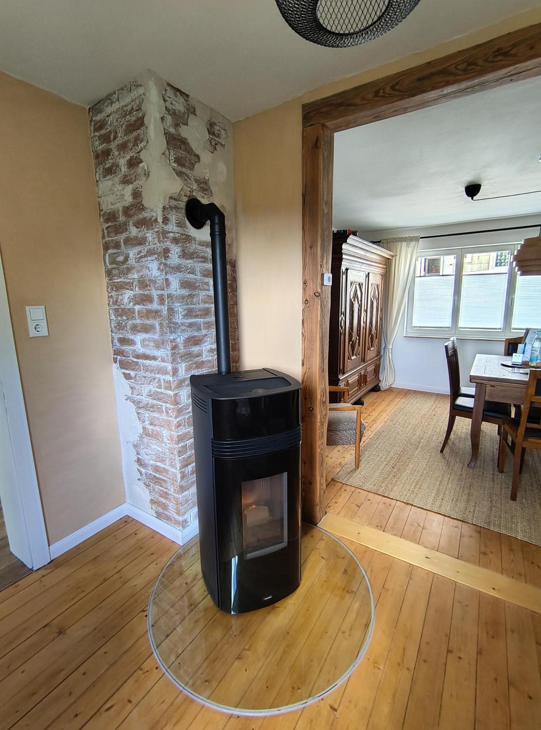 A room with a black wood-burning stove, a brick wall, wooden flooring, a dining table with chairs, and a window with blinds.