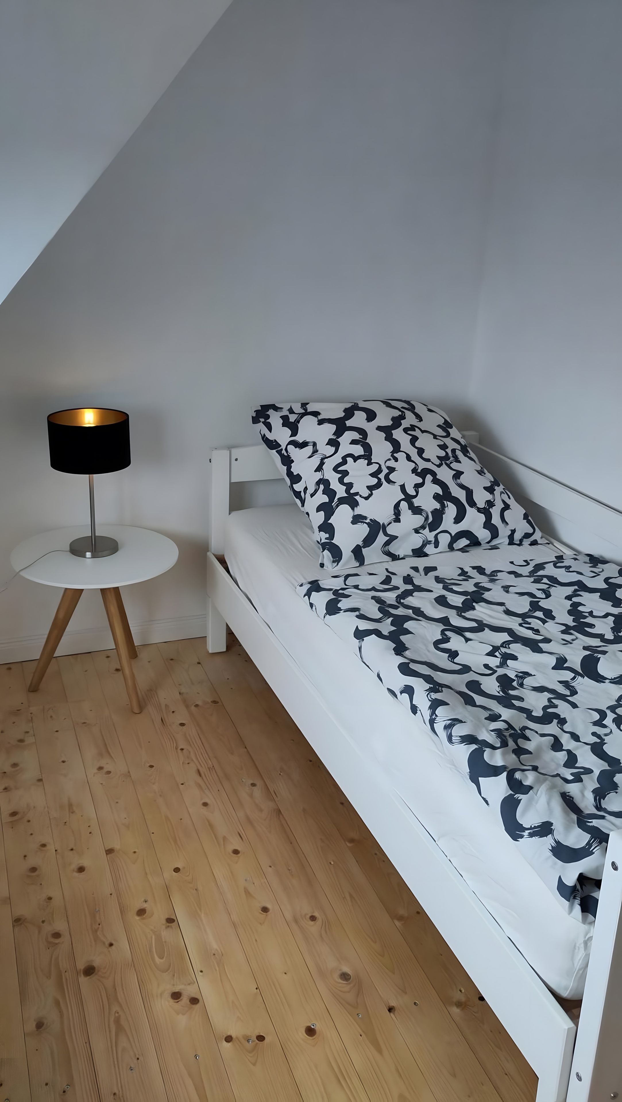 Habitación pequeña y blanca con una cama individual, techo inclinado, mesita redonda con lámpara y colcha estampada.