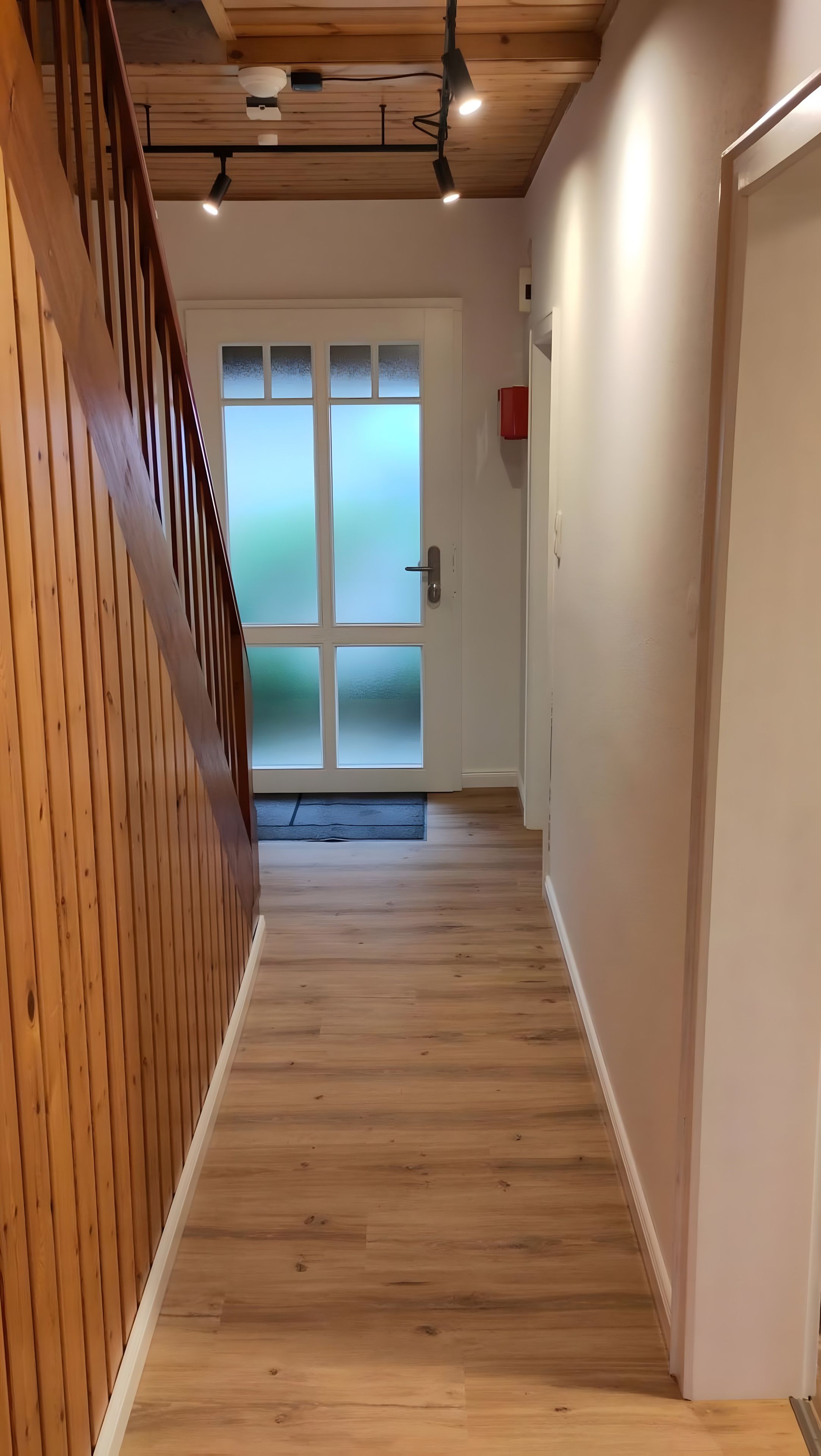 Una escalera de madera que lleva a una habitación con una puerta de vidrio esmerilado, un buzón rojo y un ventilador de techo.