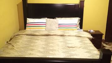 A bed with a dark headboard and a patterned bedspread, a bedside table with a lamp, and a wall with a geometric patterned floor.