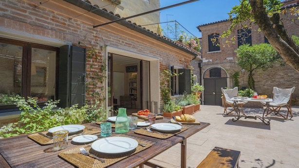 An outdoor dining area with a brick house, a table set for two, and a garden with plants and a tree. - STUNNING DORSODURO HOME WITH SECRET GARDEN & TWO PRIVATE TERRACES (Venezia)