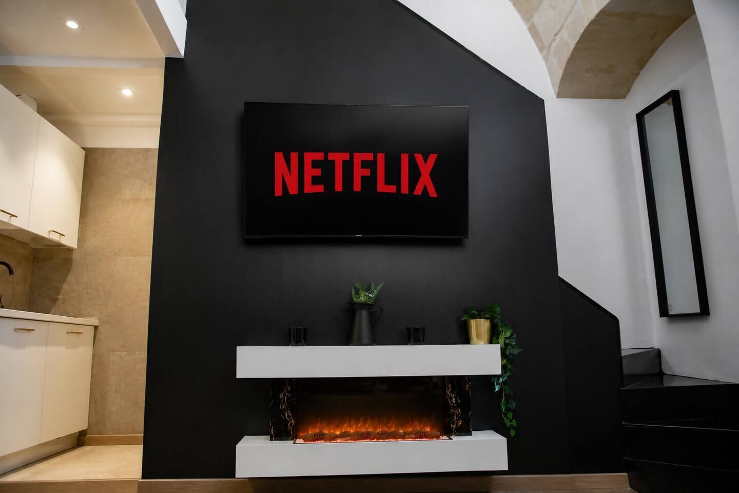 Ein modernes Wohnzimmer mit Kamin, einem an der Wand befestigten Fernseher, der 'NETFLIX' zeigt, und einer Küchenzeile links.