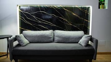 Ein modernes Wohnzimmer mit einem grauen Sofa, zwei grauen Kissen und einer Wand mit einem schwarzen und goldenen abstrakten Design.