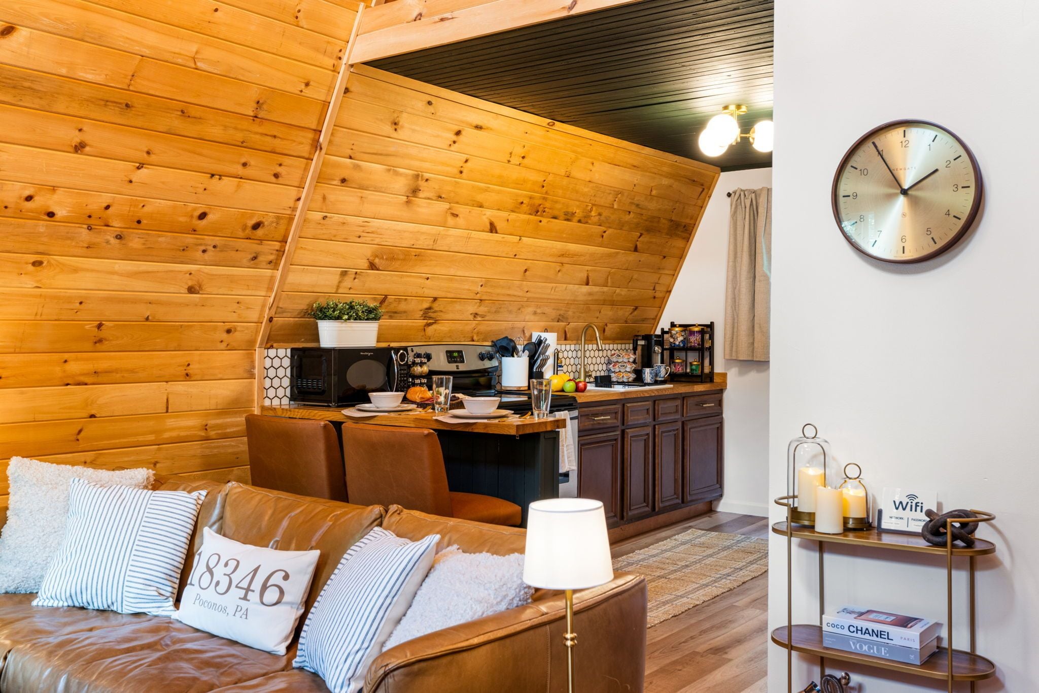 A cozy living room with a wooden ceiling, a sofa with decorative pillows, a coffee table, and a kitchen area with a microwave and toaster.