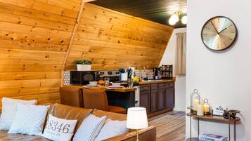 A cozy living room with a wooden ceiling, a sofa with decorative pillows, a coffee table, and a kitchen area with a microwave and toaster.