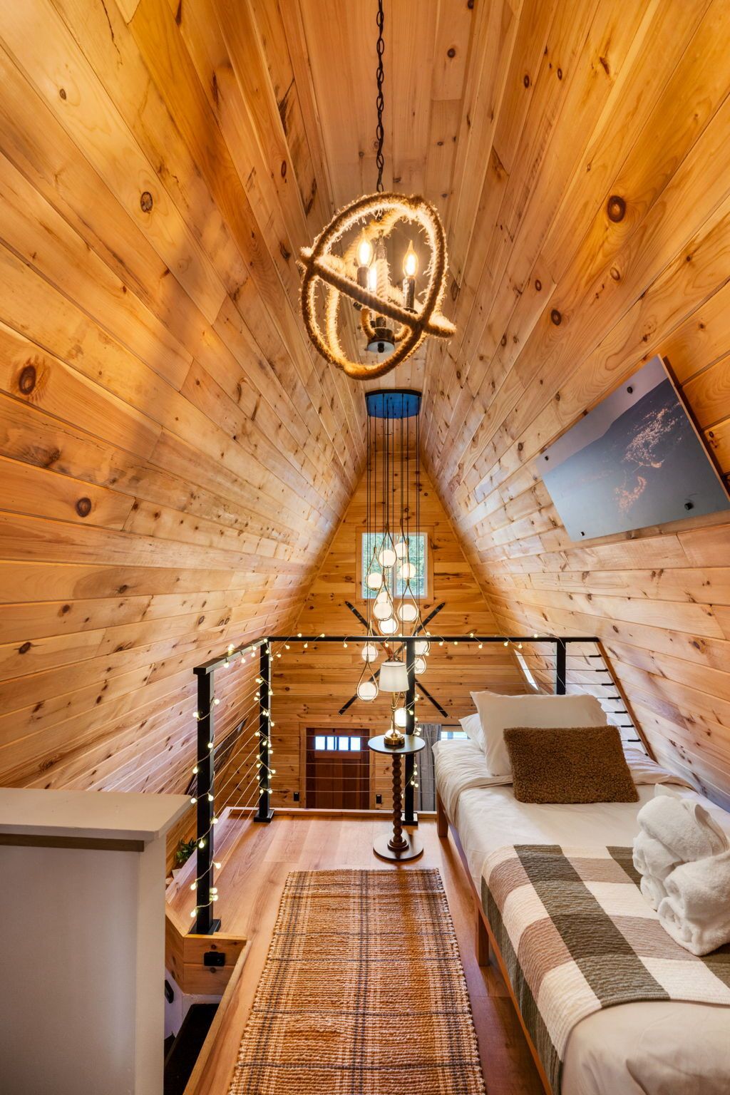 Un dormitorio acogedor en un ático con paredes de madera, una cama con almohadas, una lámpara colgante y una ventana.