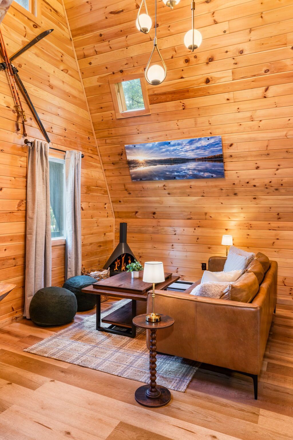 A cozy cabin interior with a wooden ceiling, a fireplace, a sofa, and a table with a lamp.