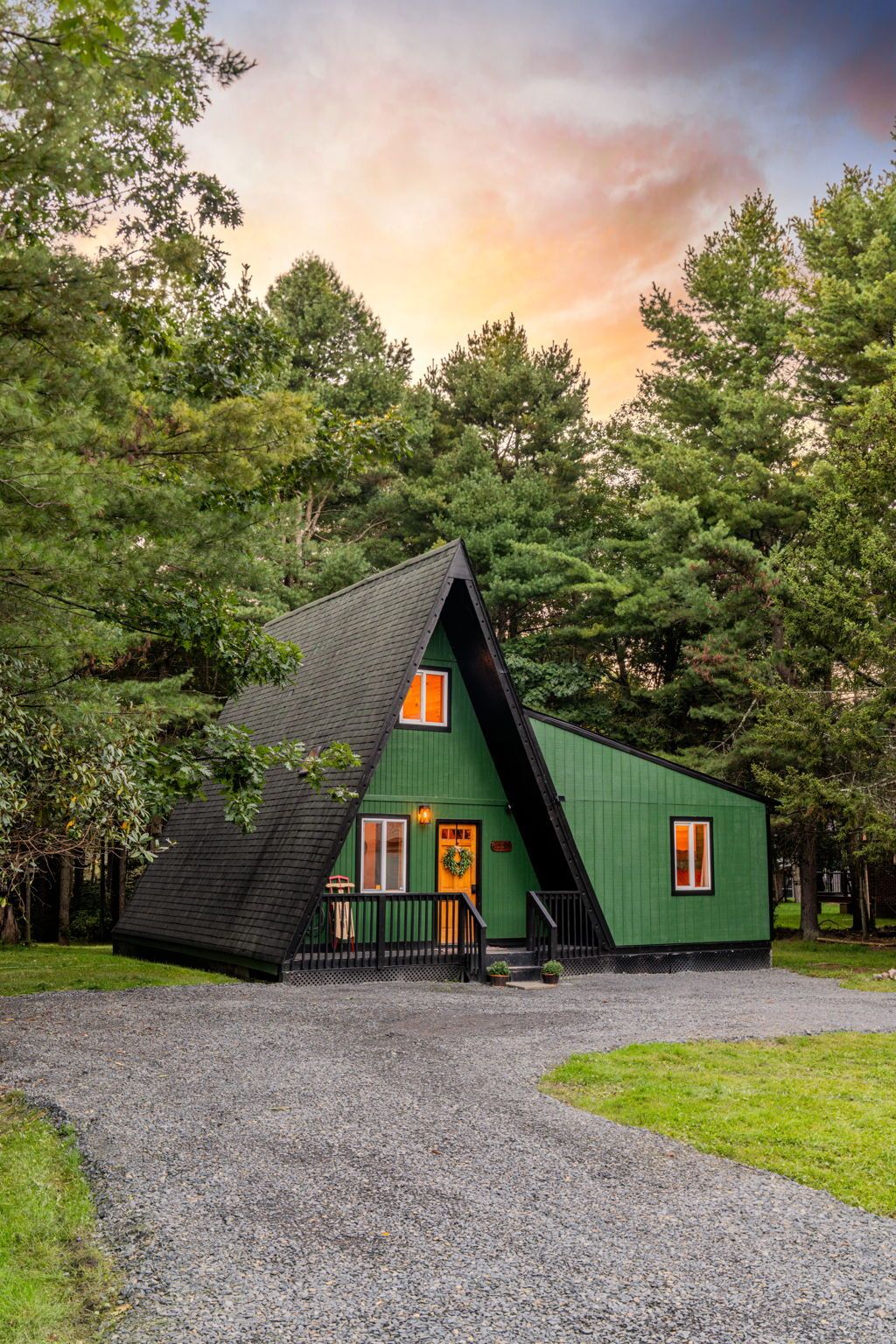 Une cabane verte, avec un toit triangulaire, située sur un chemin d’accès en gravier et entourée d’arbres.