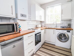 Une cuisine moderne avec des meubles blancs, un four à micro-ondes, un lave-vaisselle, une machine à laver et une fenêtre avec des stores.
