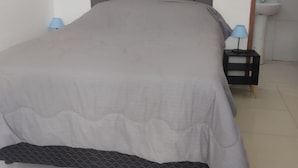 A bed with a grey headboard and a grey bedspread in a room with a white floor and a small bedside table with a lamp. - Off Road House (Ibicoara)