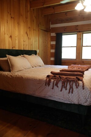 Ein Bett mit karierter Bettwäsche und einer braunen Decke, Holzwände und ein Fenster mit Vorhängen.
