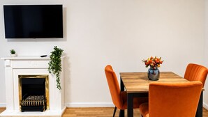 Moderni ruokailutila, jossa on puinen pöytä, oranssit tuolit, seinälle asennettu litteänäyttöinen televisio takan yläpuolella ja pieni kasvi takan reunalla.