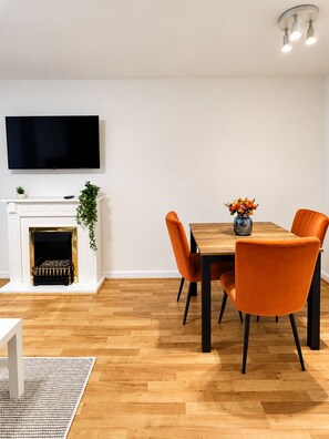 Ein moderner Essbereich mit einem Holztisch, orangefarbenen Stühlen, einem flachbildfernseher über einem Kamin und einer kleinen Pflanze auf dem Sims.