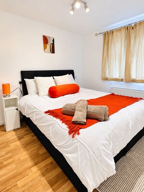 Ein ordentlich gemachtes Bett mit weißer Bettwäsche und einer roten Tagesdecke, ein Nachttisch mit einer Lampe und ein Fenster mit Vorhängen.