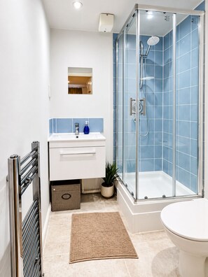 Ein modernes Badezimmer mit einer gläsernen Duschkabine, einem weißen Waschbecken und einem blauen Seifenspender sowie einer beigefarbenen Fußmatte.