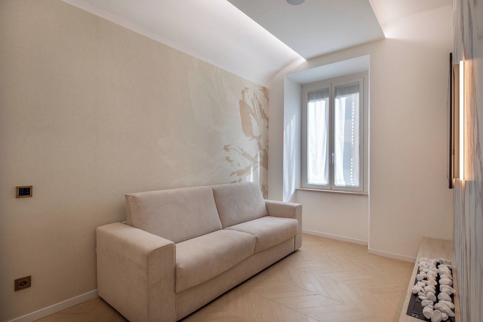 Un salon minimaliste avec un canapé beige, un parquet en bois à motifs chevrons, une grande fenêtre avec des rideaux légers et un luminaire fixé au mur.